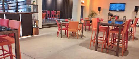 A spacious indoor area with multiple seating arrangements, including a bar area with a counter and chairs, and a dining area with tables and chairs. There are ceiling fans and a television mounted on the wall.