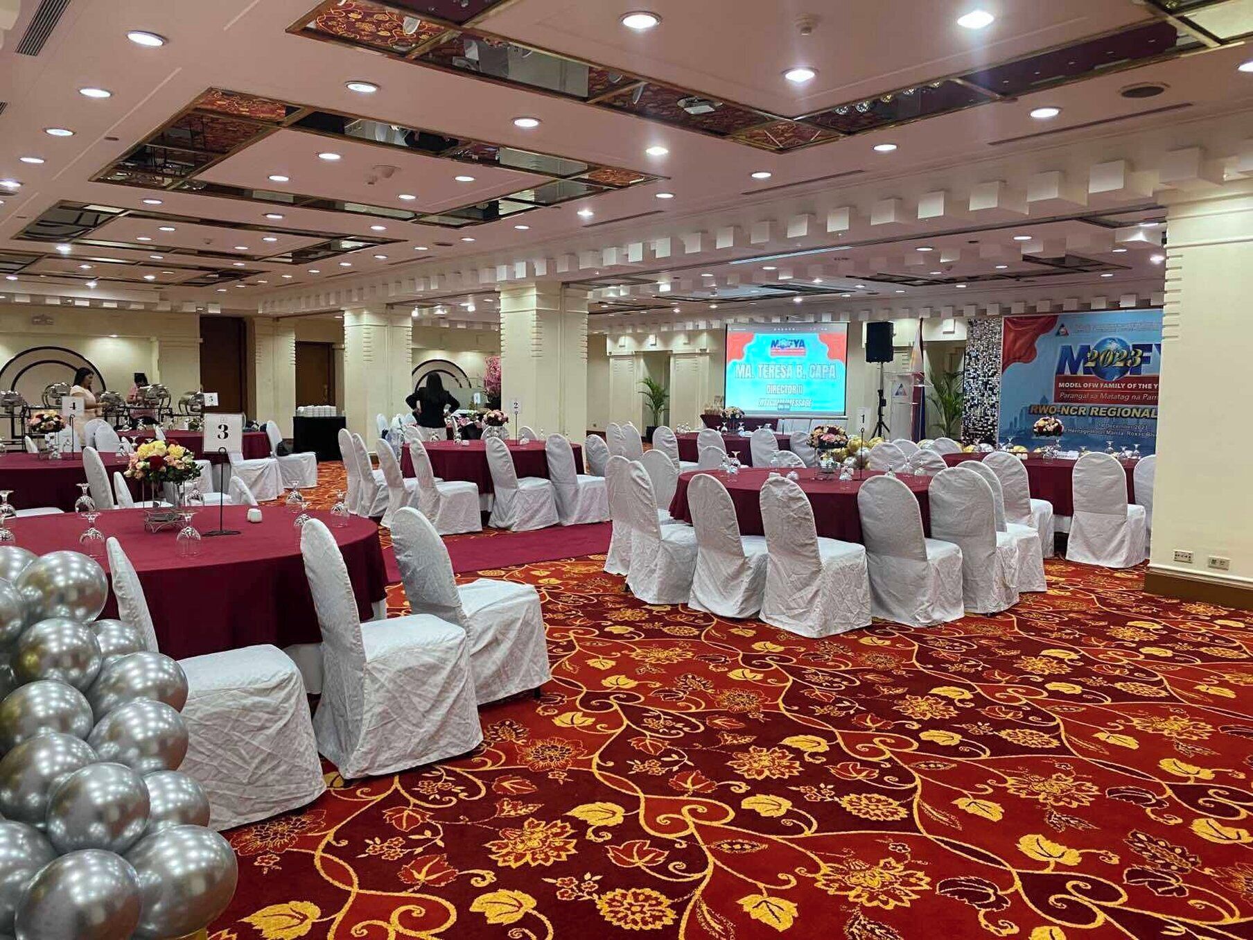 A banquet hall with round tables covered in red tablecloths and white chair covers, arranged in rows.