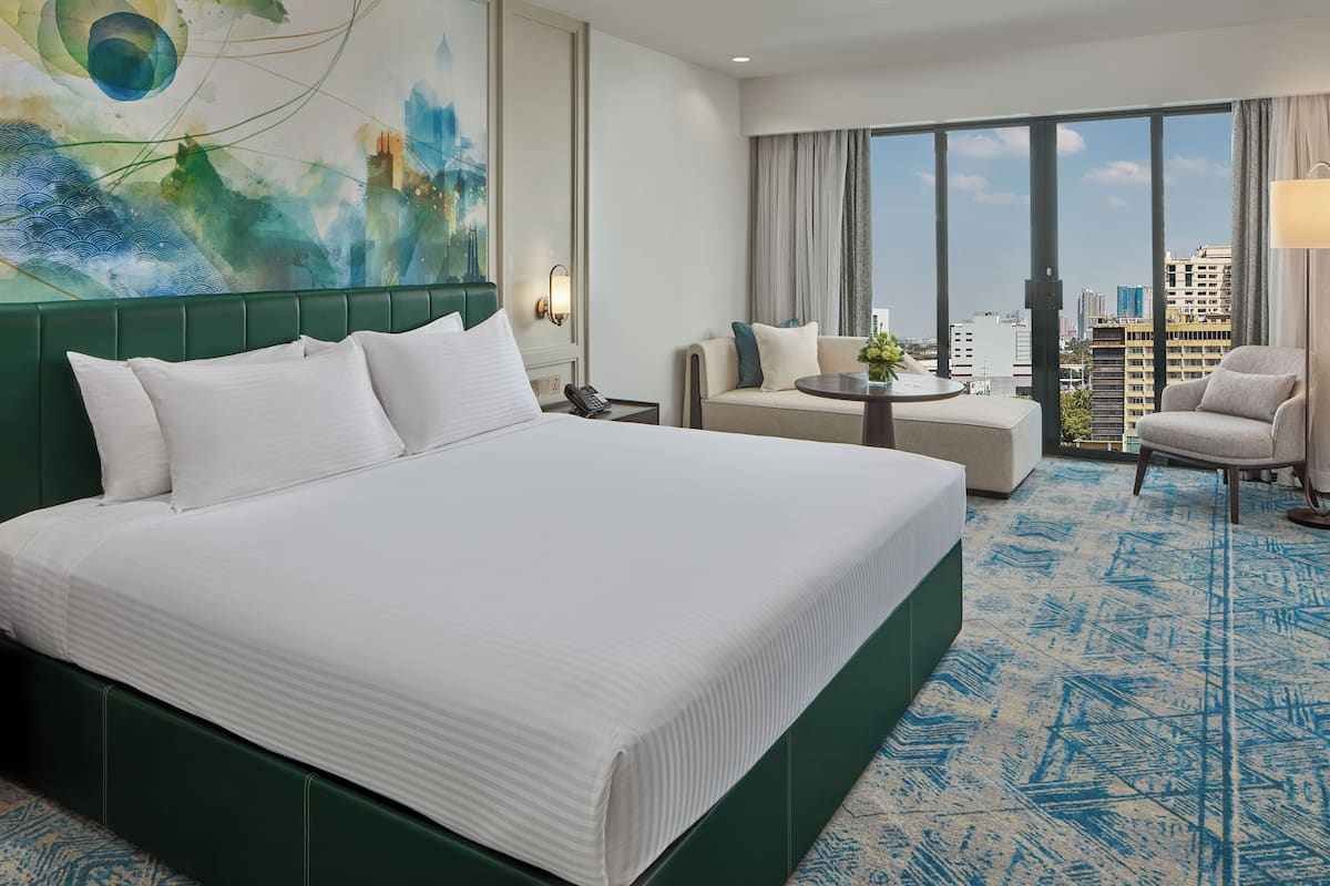 A hotel room with a large bed, a sofa, a small table, and a chair. There is a city view through the windows.