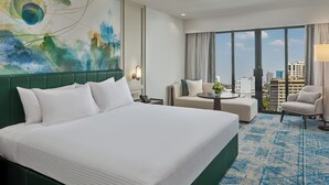 Habitación de hotel con una cama grande, un sofá, una mesita y una silla. Se aprecia vista de la ciudad a través de las ventanas.
