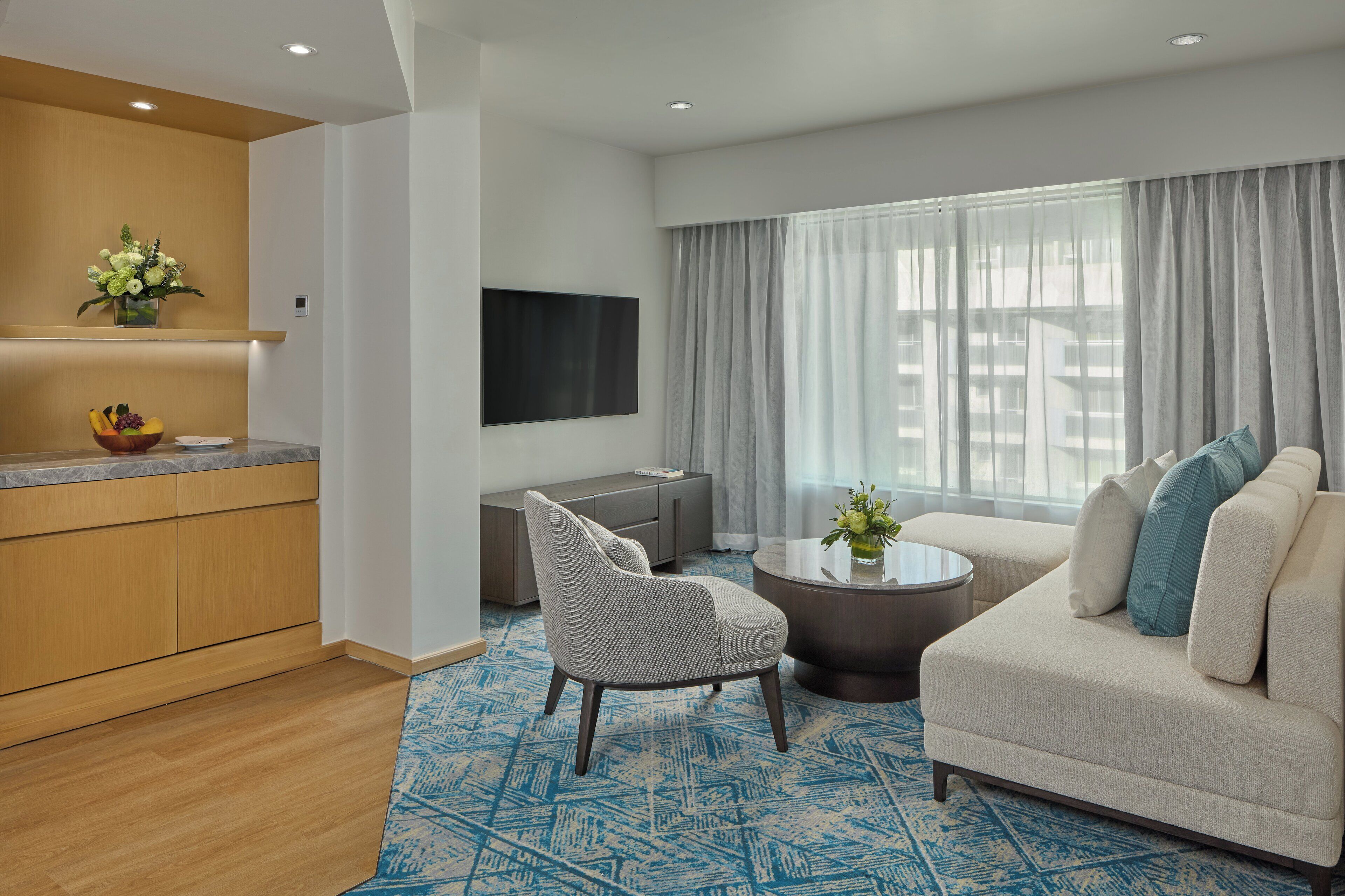 A modern hotel room with a living area, a kitchen, and a large window with curtains.
