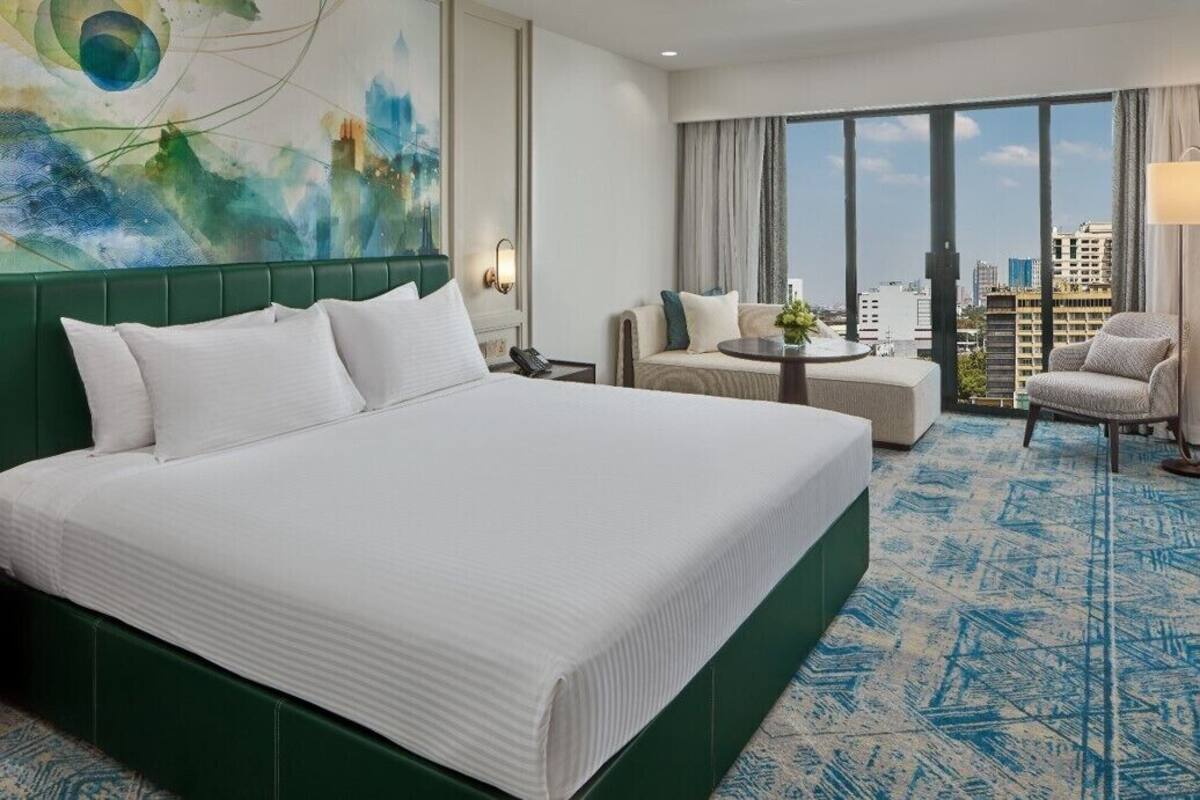A hotel room with a large bed, a sofa, a small table, and a view of the city through large windows.