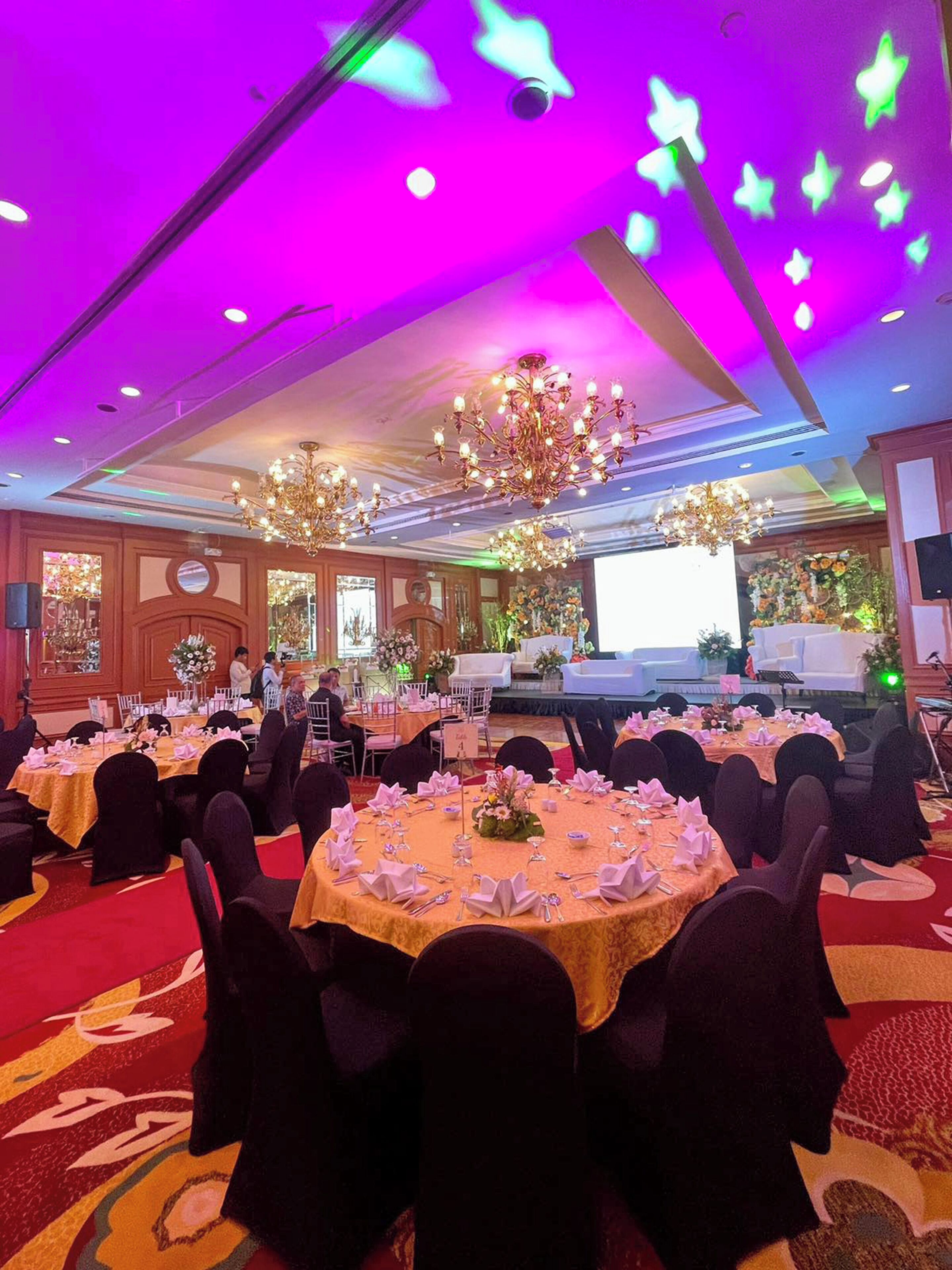 Un salón de banquetes con mesas redondas dispuestas para comer, candelabros y un escenario para presentaciones.
