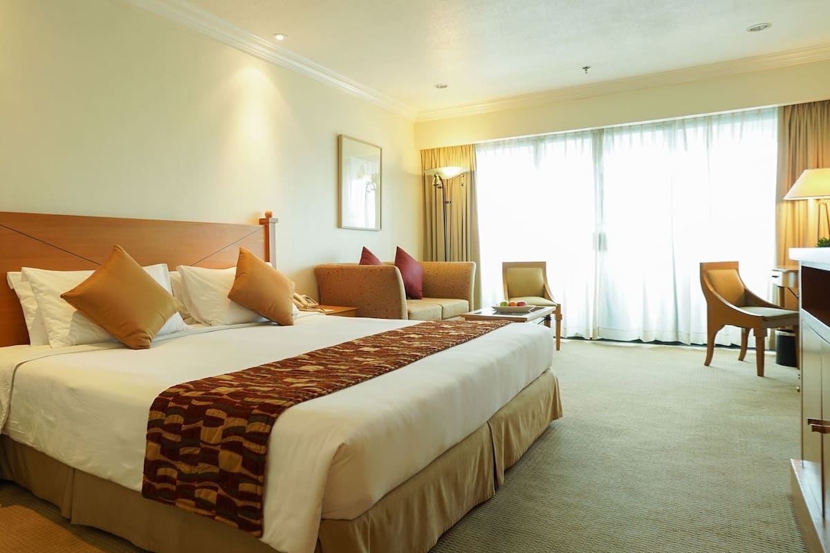 A hotel room with a large bed, a desk, two chairs, a lamp, and a window with curtains.