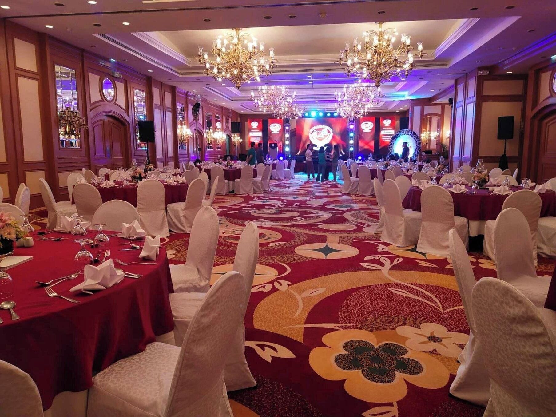 A banquet hall with round tables, white chairs, chandeliers, and a stage with a large screen.