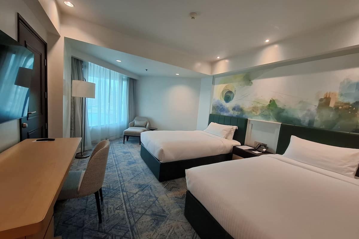 A hotel room with two beds, a desk, a chair, a lamp, and a painting on the wall.