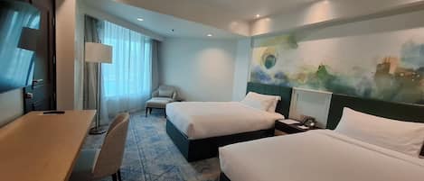 A hotel room with two beds, a desk, a chair, a lamp, and a painting on the wall.