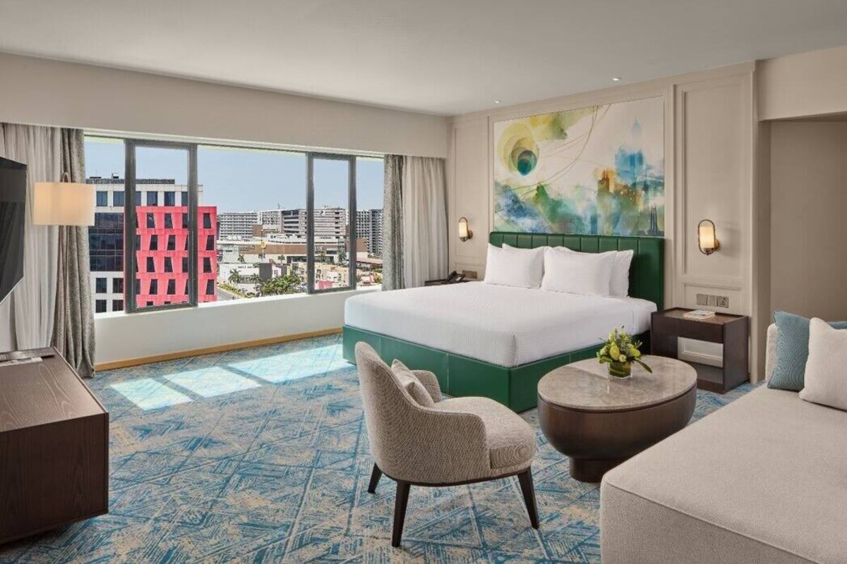 A hotel room with a large bed, a chair, a round coffee table, and a view of the city through large windows.