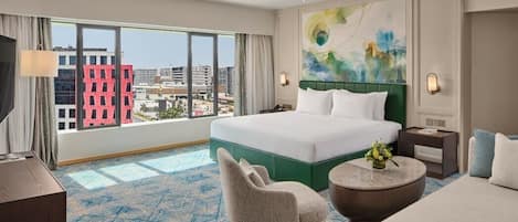Une chambre d’hôtel avec un grand lit, une chaise, une table basse ronde et une vue sur la ville grâce à de grandes fenêtres.