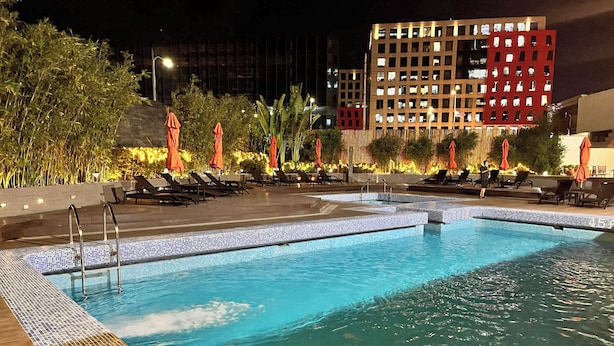 Un área de piscina nocturna con sillones de descanso y sombrillas, rodeada de vegetación y un edificio moderno al fondo.