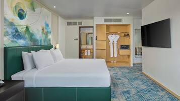 A hotel room with a large bed, a green headboard, a flat-screen TV, a wardrobe, and a bathroom visible through an open door.