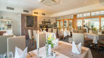 L’intérieur d’un restaurant, avec des tables dressées pour le service, un menu écrit à la craie et une vue dégagée grâce à de grandes fenêtres.