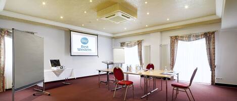 Un salón de conferencias con una pantalla de proyector mostrando 'Best Western', una pizarra, una mesa con sillas y suelo alfombrado.
