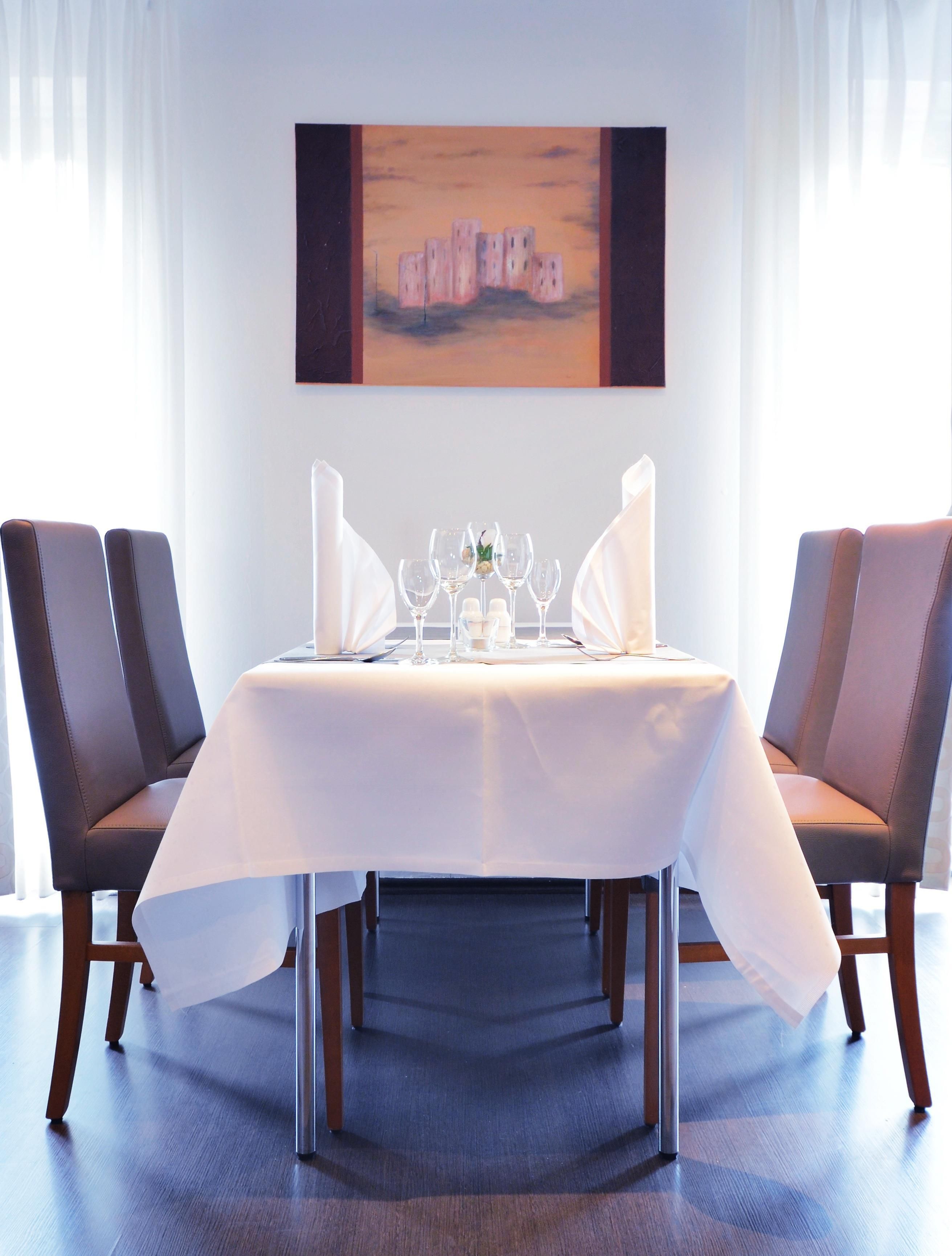 Une table à manger recouverte d’une nappe blanche, entourée de quatre chaises, dans une pièce où un tableau est accroché au mur.
