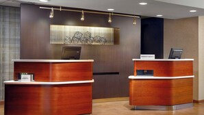 A hotel reception area with two wooden reception desks, a modern wall art piece, and a nameplate on the right desk. - Courtyard by Marriott Fayetteville (Fayetteville)