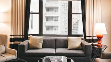 A modern living room with a grey sofa, two cushions, a glass coffee table, and a view of a cityscape through large windows.