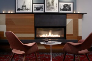 Una habitación con chimenea, dos sillones, una mesa con un libro y una copa de vino, y cuadros enmarcados sobre la chimenea.