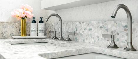 Un baño moderno con encimera de granito, un lavamanos y dos grifos. Hay dos botellas de loción y un florero con flores sobre la encimera.