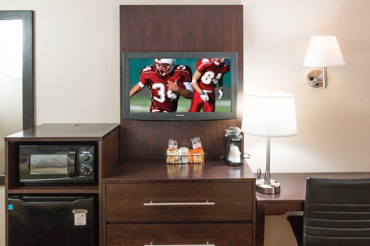 A hotel room with a TV displaying a football game, a microwave, a mini-fridge, a lamp, and a chair.