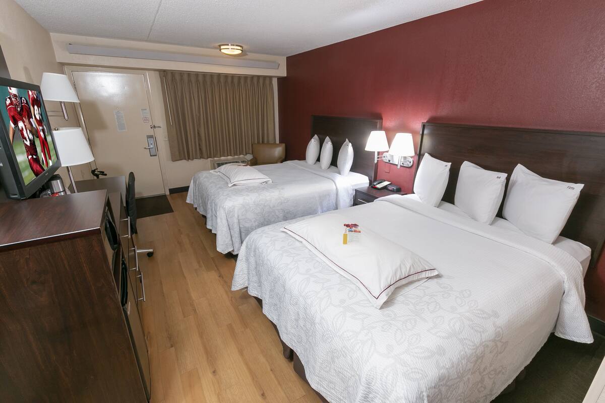 Habitación de hotel con dos camas, un escritorio con televisor y un baño visible a través de una puerta.