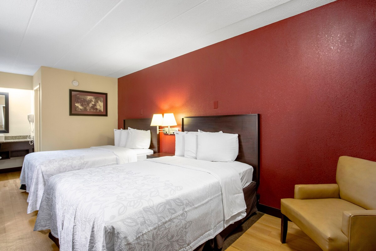 A hotel room with two beds, a red accent wall, a wooden floor, a chair, and a nightstand with a lamp.