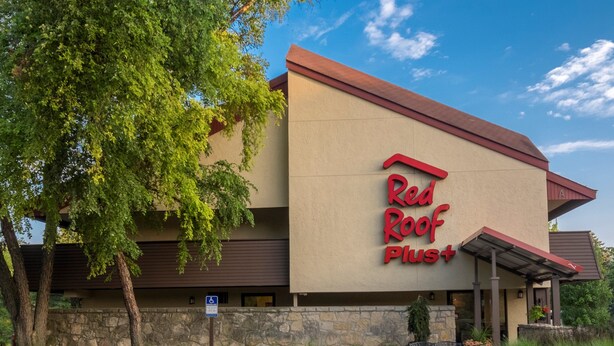 Un hôtel Red Roof Plus avec une façade en pierre, un parking et un ciel d’un bleu éclatant.