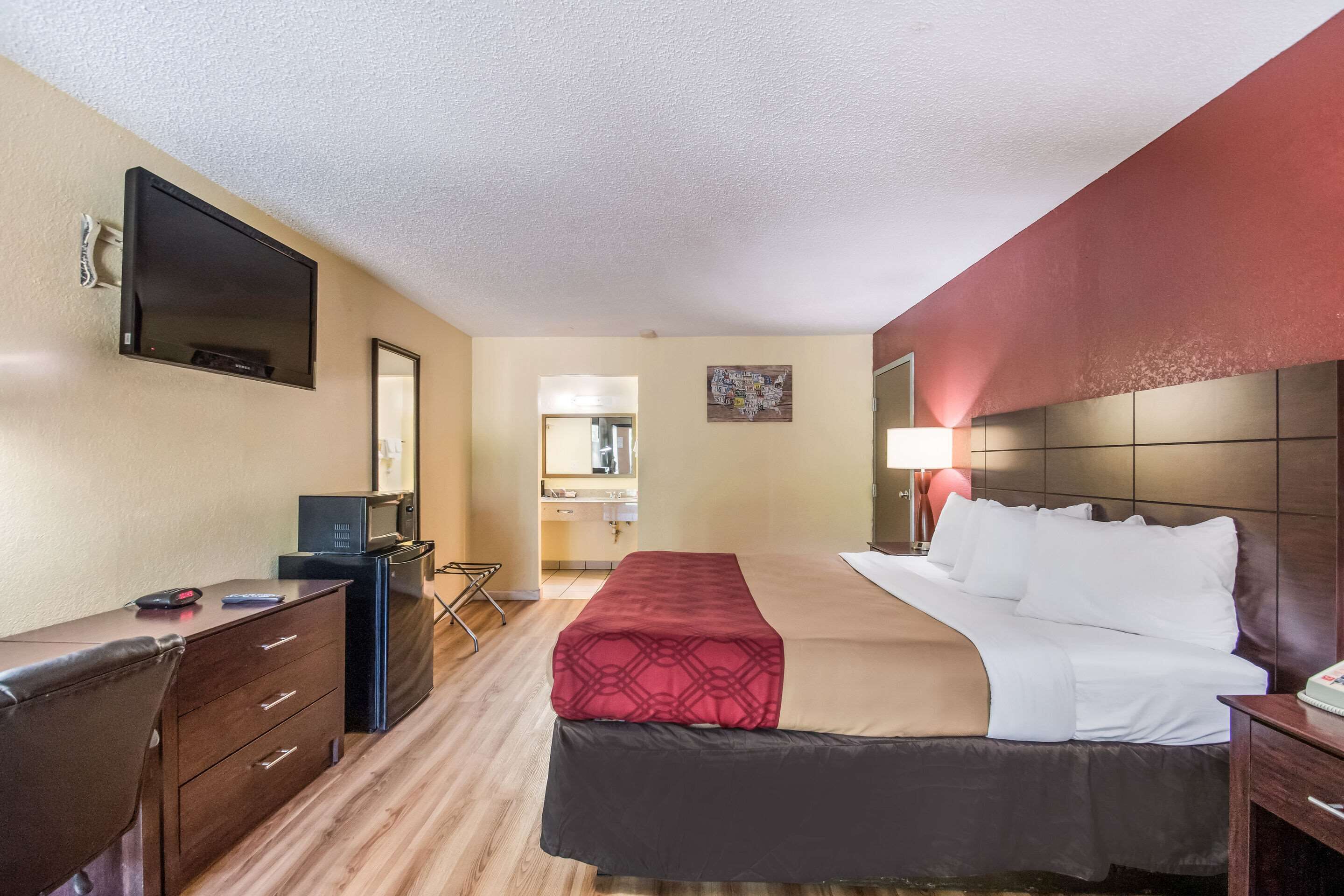A hotel room with a bed, a desk with a computer, a TV, and a bathroom visible through an open door.