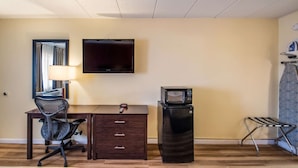 A compact hotel room with a wooden desk, a black office chair, a flat-screen TV mounted on the wall, a black refrigerator, and a small metal rack. - Econo Lodge Kearney - Liberty (Kearney)