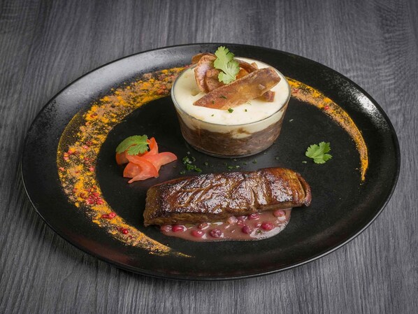 A plate with a grilled meat, a layered dessert, and garnishes on a dark surface.