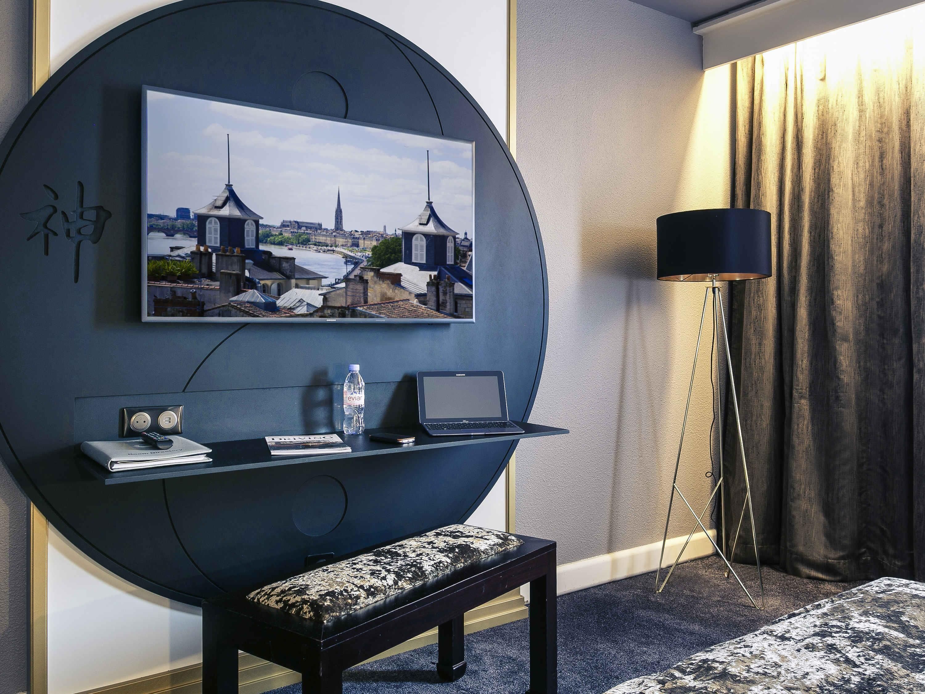 Una habitación de hotel moderna con un televisor redondo grande montado en la pared que muestra un paisaje urbano, una computadora portátil sobre una estantería suspendida, una botella de agua, un control remoto y un banco.