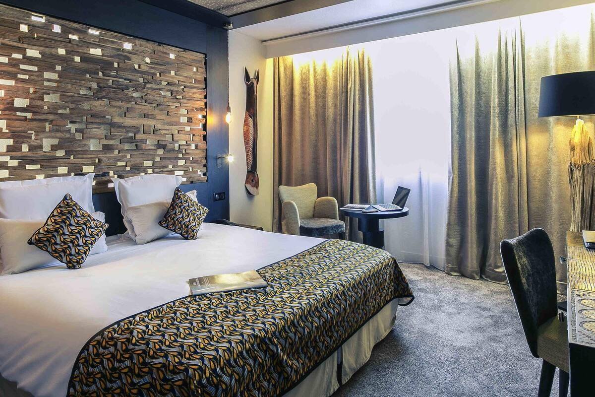 A hotel room with a large bed, a small table with a laptop, a chair, and a view of trees through the window.