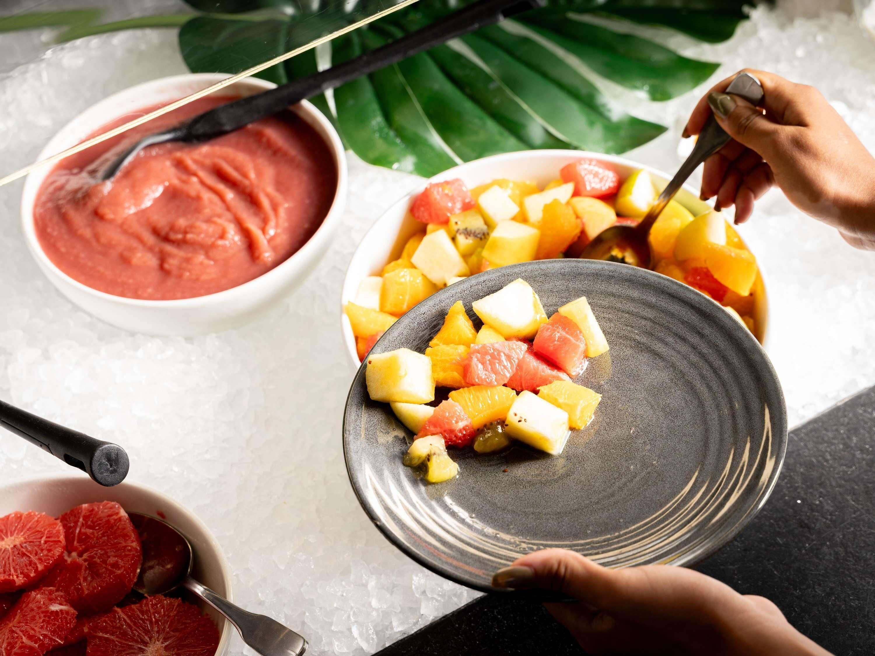 A person serving fruit onto a plate, with a bowl of pink sauce and grapefruit slices on ice nearby.