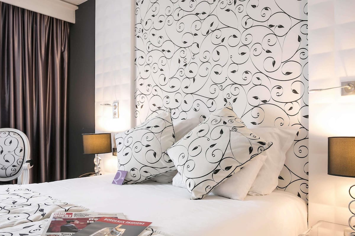 Habitación de hotel con una cama, dos lámparas de noche, una mesita de noche, una silla y una pared con estampado floral.