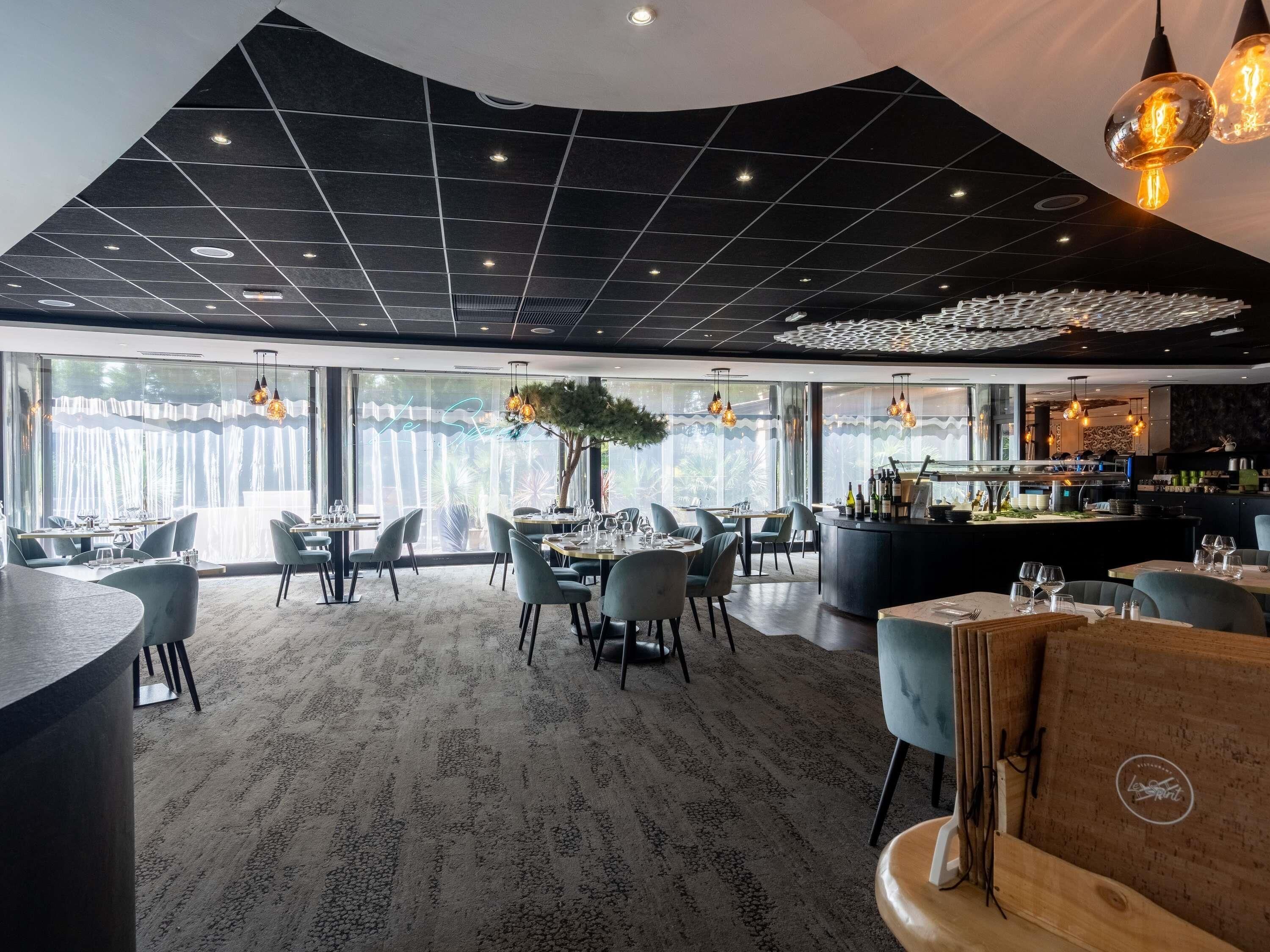 A modern restaurant interior with a high ceiling, large windows, and a central bar area.
