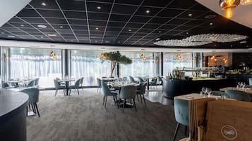 A modern restaurant interior with a high ceiling, large windows, and a central bar area.