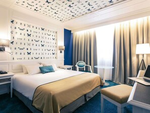 A hotel room with a bed, a desk, a chair, a lamp, and a window with curtains.