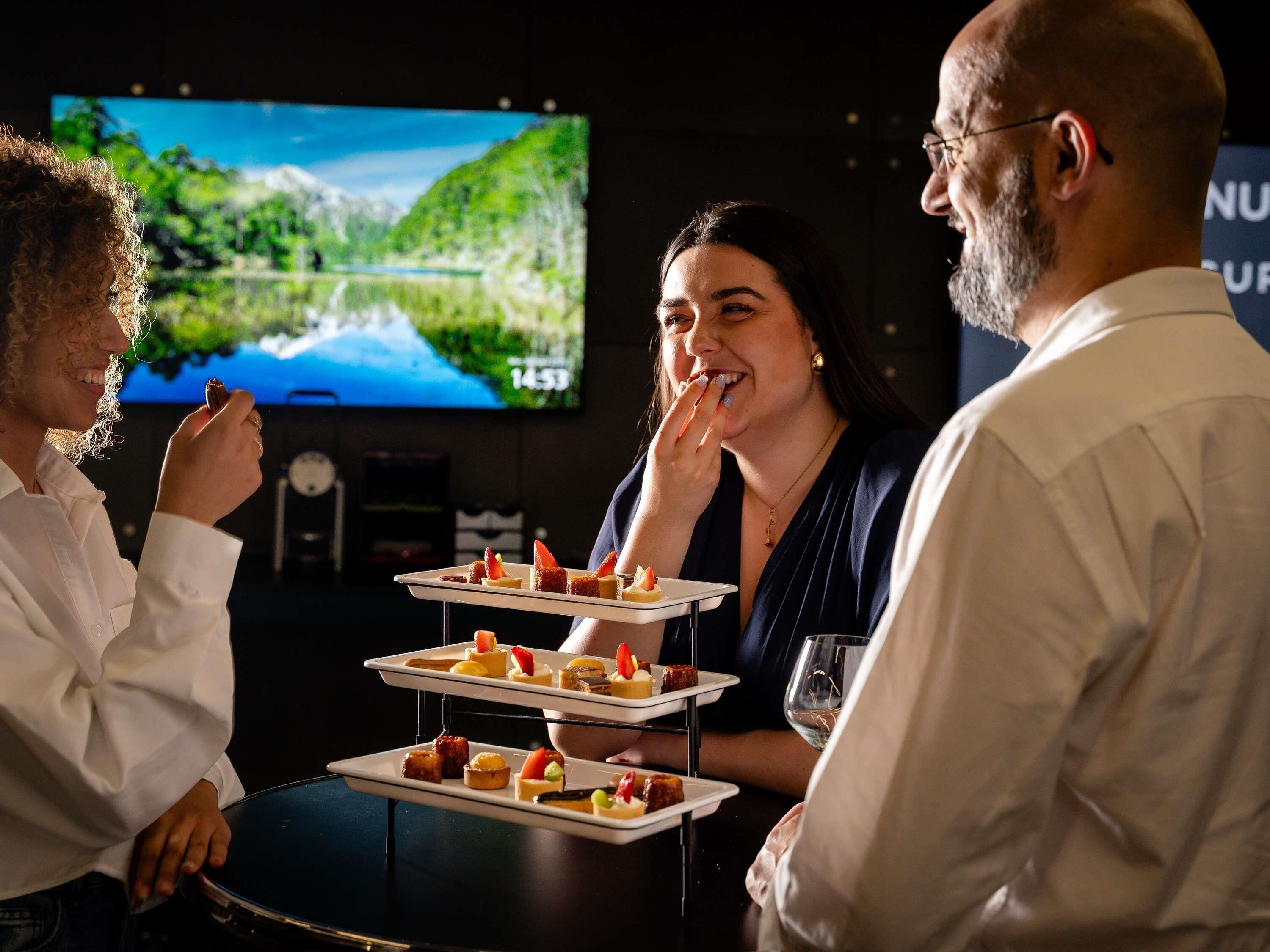 Un grupo de personas sentadas alrededor de una mesa con una bandeja de postres, una pantalla de televisión al fondo y un reloj.