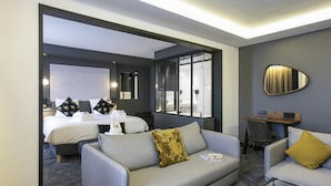 A modern hotel room with a grey sofa, a glass coffee table, and a bed with white linens and decorative pillows.