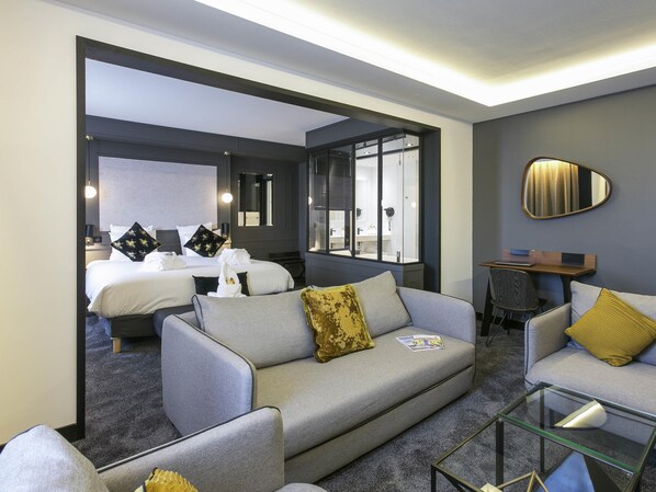 Una habitación de hotel moderna con un sofá gris, una mesa de centro de vidrio y una cama con sábanas blancas y almohadones decorativos.