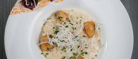 Un plato blanco con un guiso cremoso, adornado con hierbas picadas y queso rallado, acompañado de rodajas de remolacha y una guarnición crujiente.
