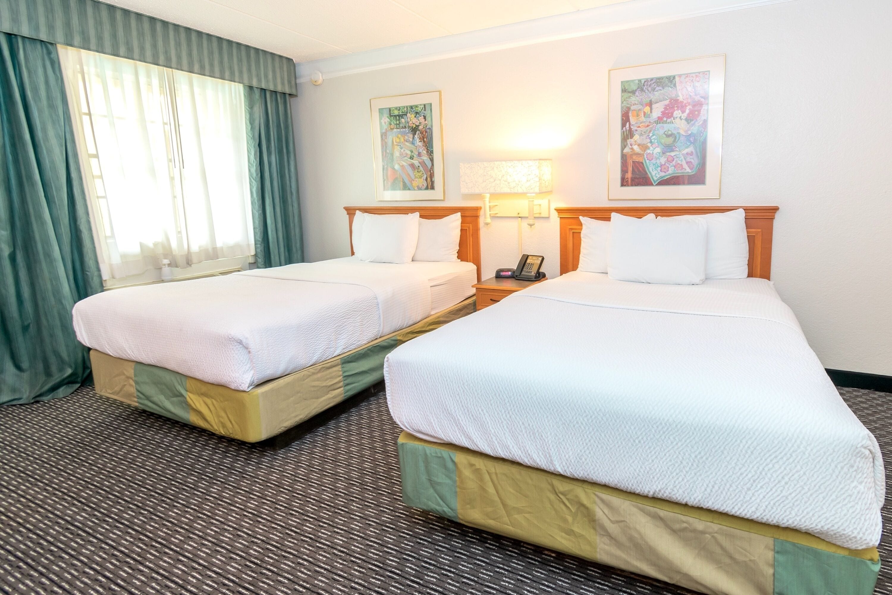 Una habitación de hotel con dos camas, una ventana con cortinas, una lámpara y dos cuadros en la pared.