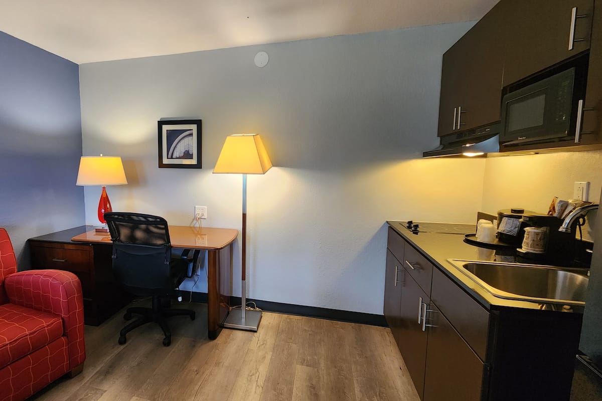 Una habitación de hotel compacta con kitchenette, un escritorio con dos lámparas, una silla roja y un cuadro enmarcado en la pared.