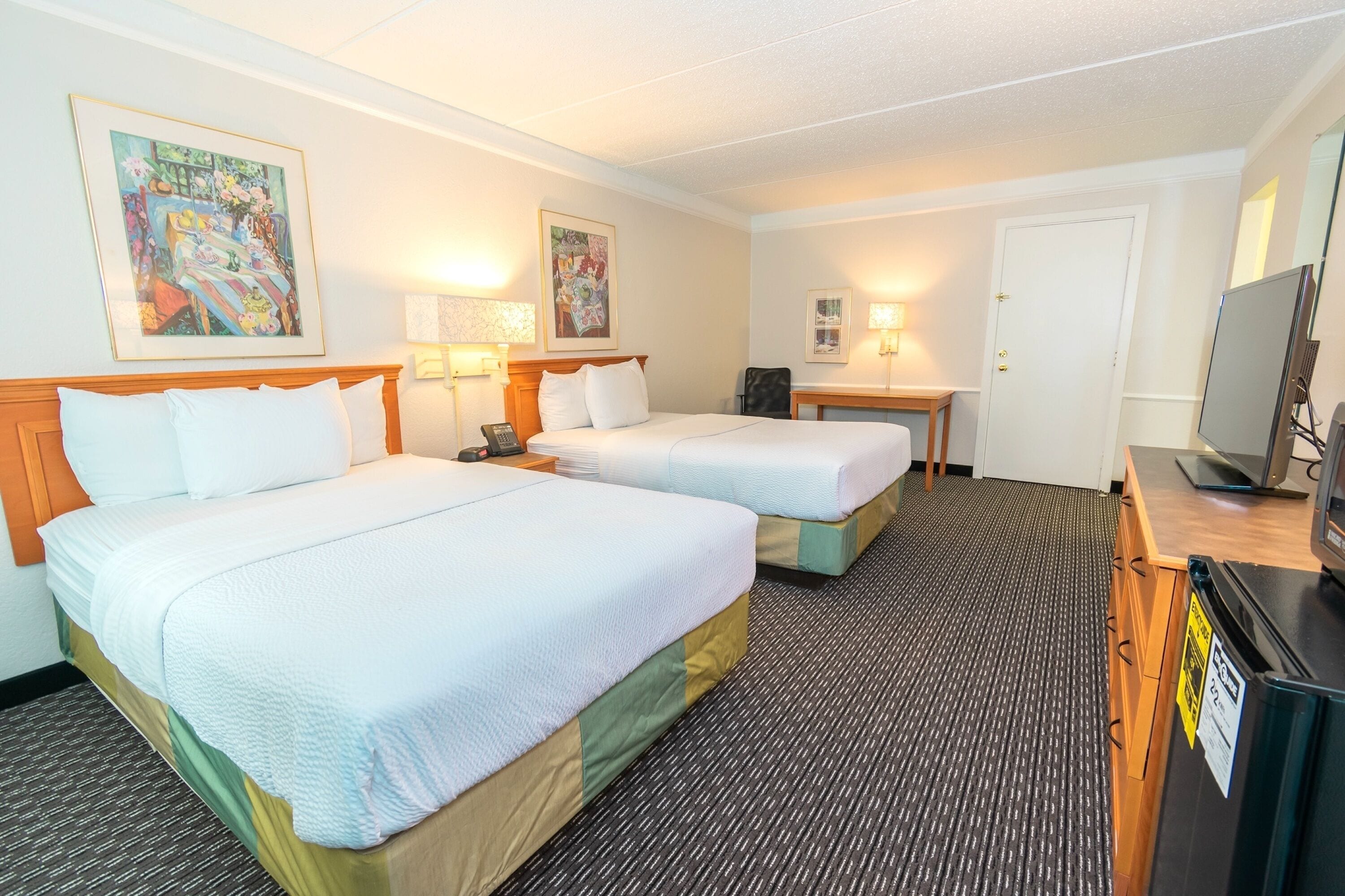 A hotel room with two beds, a desk, a chair, a television, and artwork on the walls.