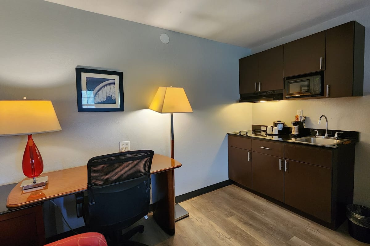 Habitación de hotel compacta con kitchenette, un escritorio con dos lámparas, una silla y un cuadro en la pared.