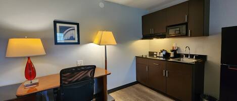 A compact hotel room with a kitchenette, a desk with two lamps, a chair, and a framed picture on the wall.