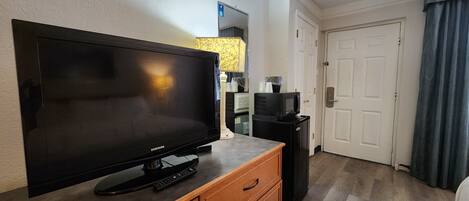 A hotel room with a large flat-screen TV on a wooden cabinet, a microwave, a small refrigerator, and a door with a keypad lock.