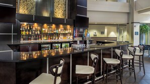A bar area with a long counter, multiple stools, and a variety of bottles. - Embassy Suites by Hilton St. Louis Airport (Bridgeton)