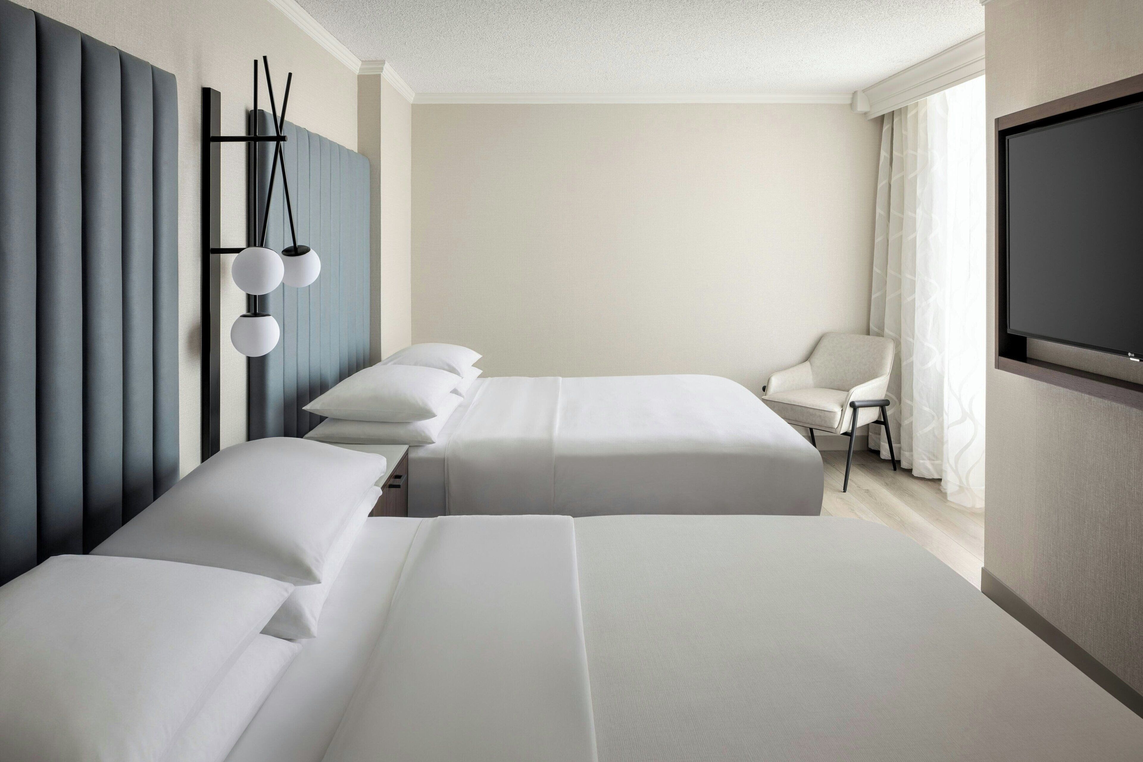A hotel room with two beds, a TV, a chair, and a wall-mounted light fixture.