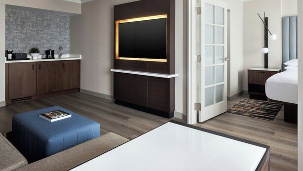 A modern hotel room with a flat-screen TV, a sofa, a coffee table, a small kitchen area, and a separate bedroom with a bed and nightstand. - Chicago Marriott Suites Deerfield (Deerfield)
