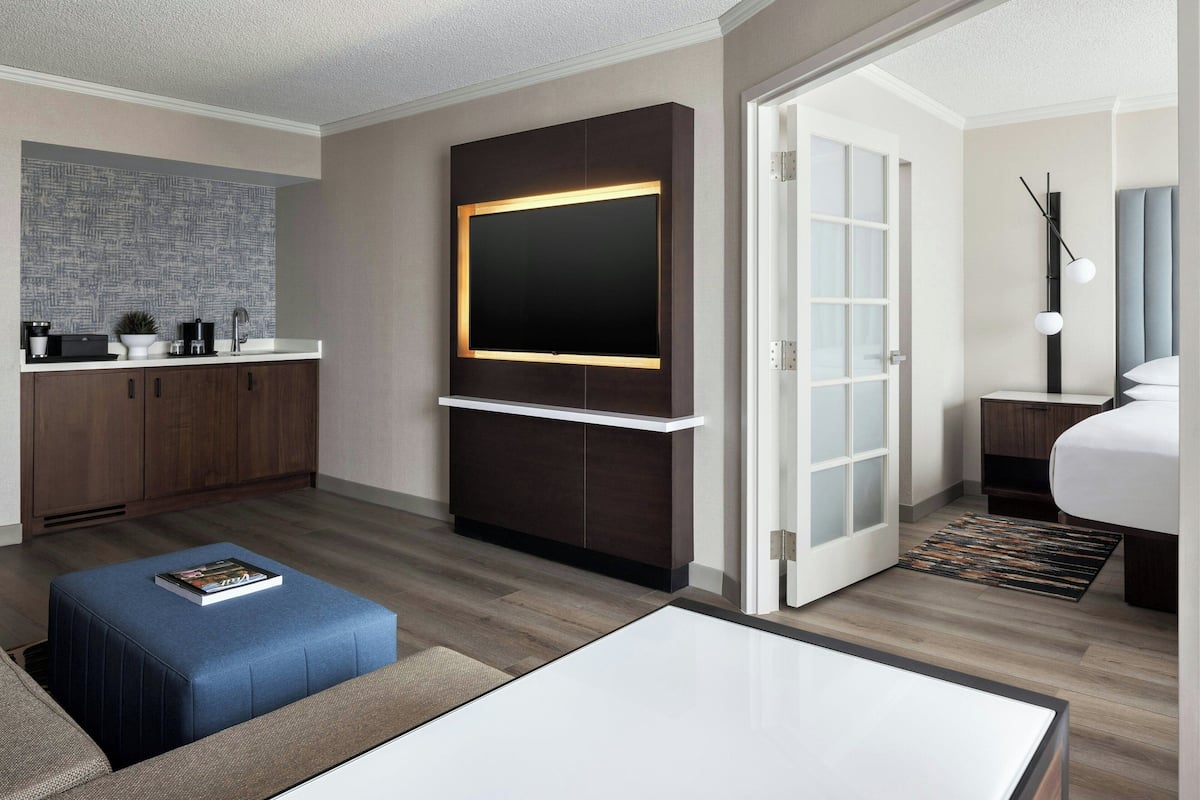 Habitació d'hotel moderna amb televisió de pantalla plana, sofà, taula de cafè, zona de cuina petita i dormitori separat amb llit i nitòri.